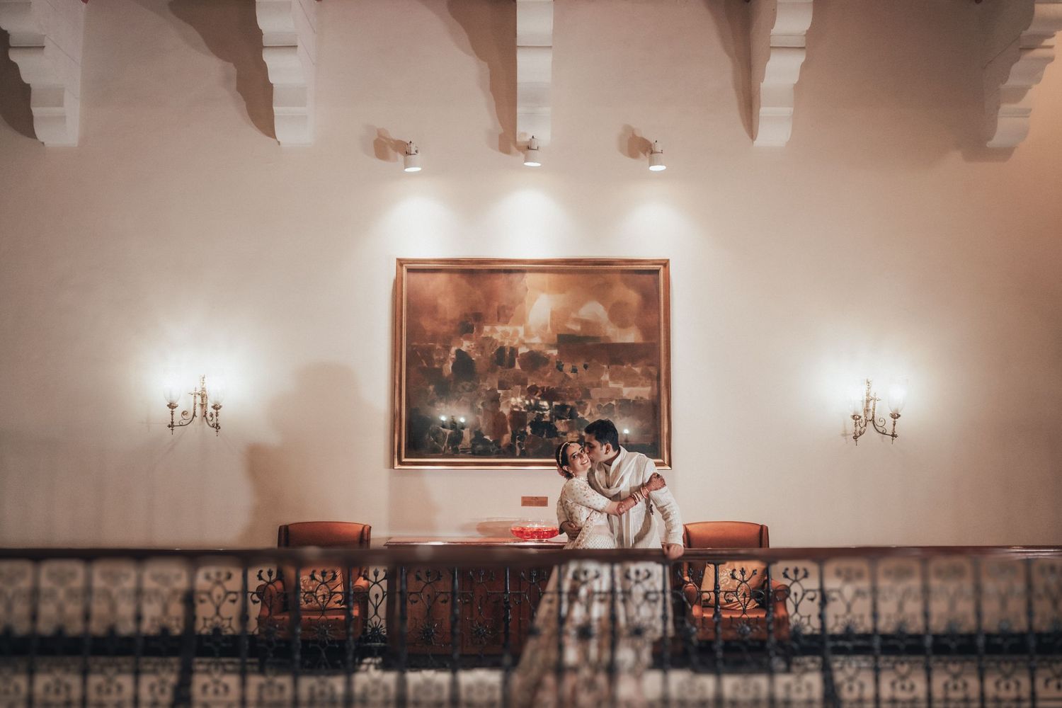 Indian wedding couple shares intimate embrace beneath large painting in warmly lit elegant venue.