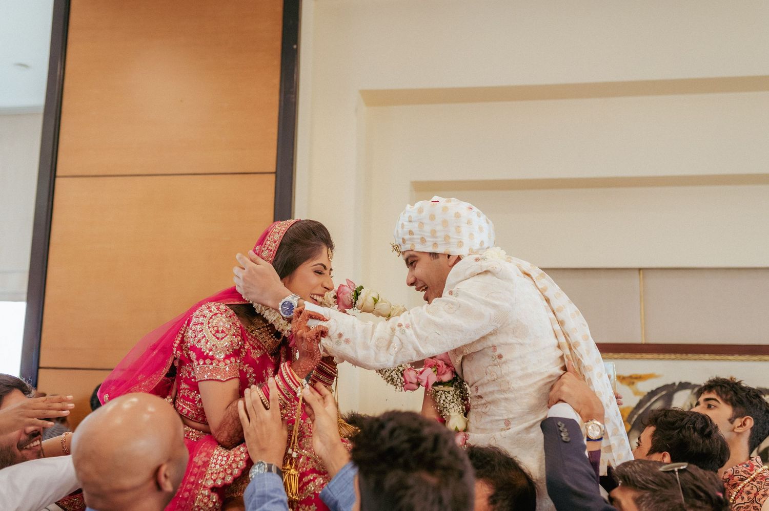 Indian bride and groom in traditional attire exchange garlands surrounded by guests at wedding.