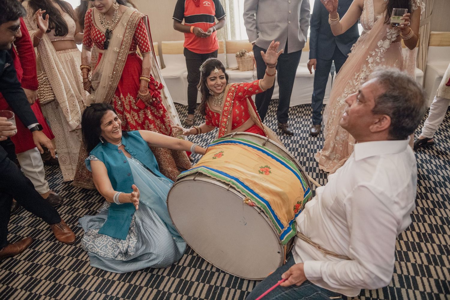 Guests playing dhol drum together on floor during vibrant Indian wedding celebration.