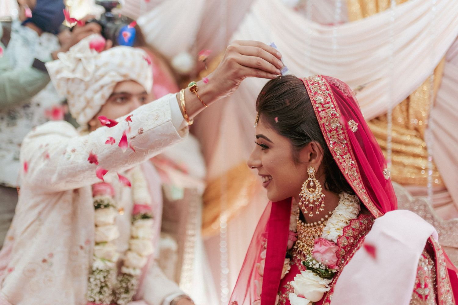 Groom applies sindoor to bride's hair parting during colorful outdoor Hindu wedding ceremony with flower petals.