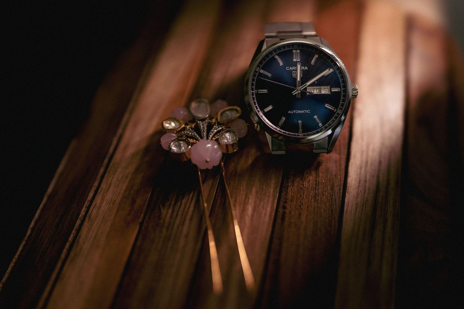A classic blue-dial wristwatch beside delicate pink floral brooches on a wooden surface.