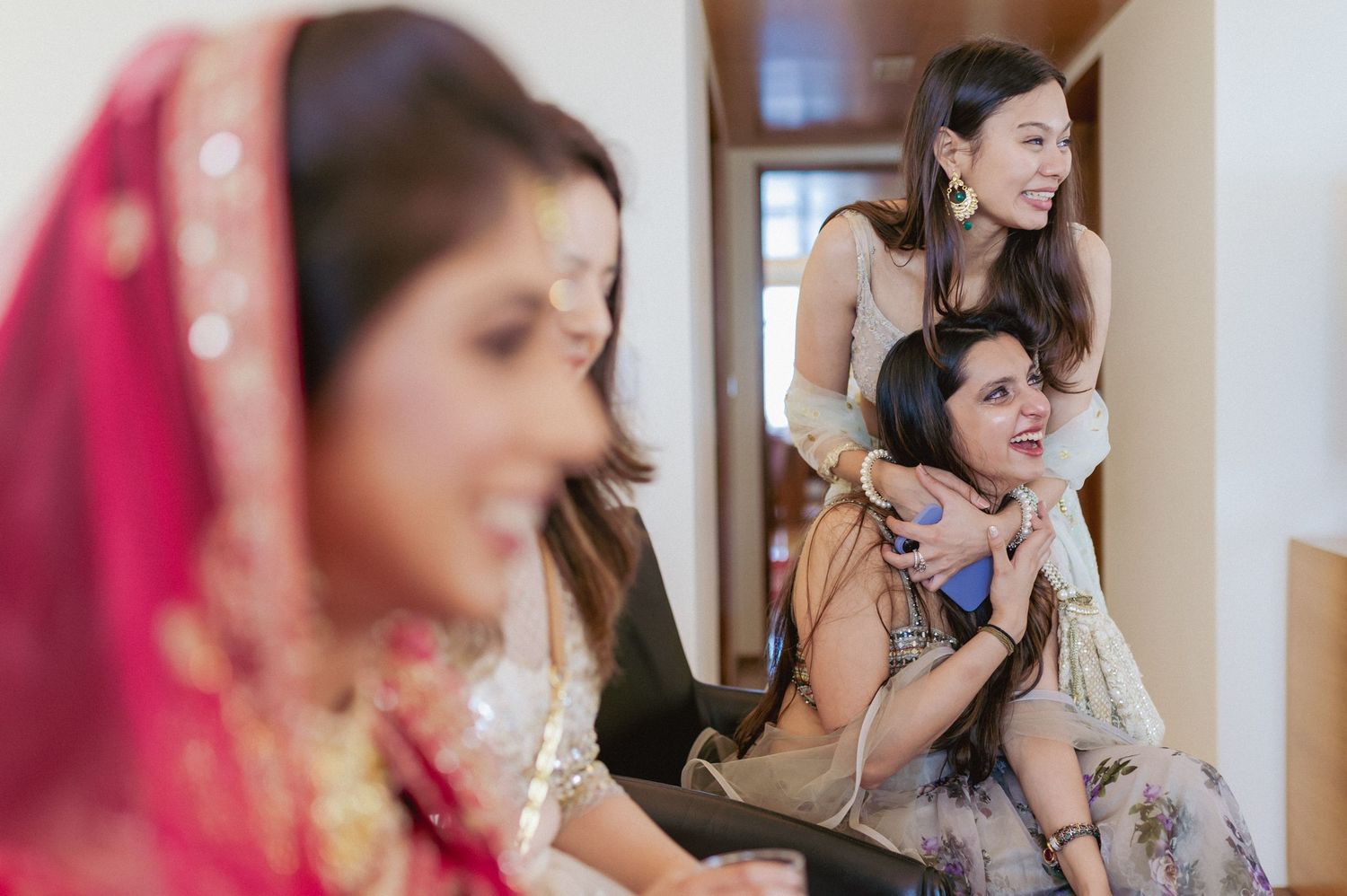 Two women laugh and embrace in hallway while Indian bride stands in foreground.