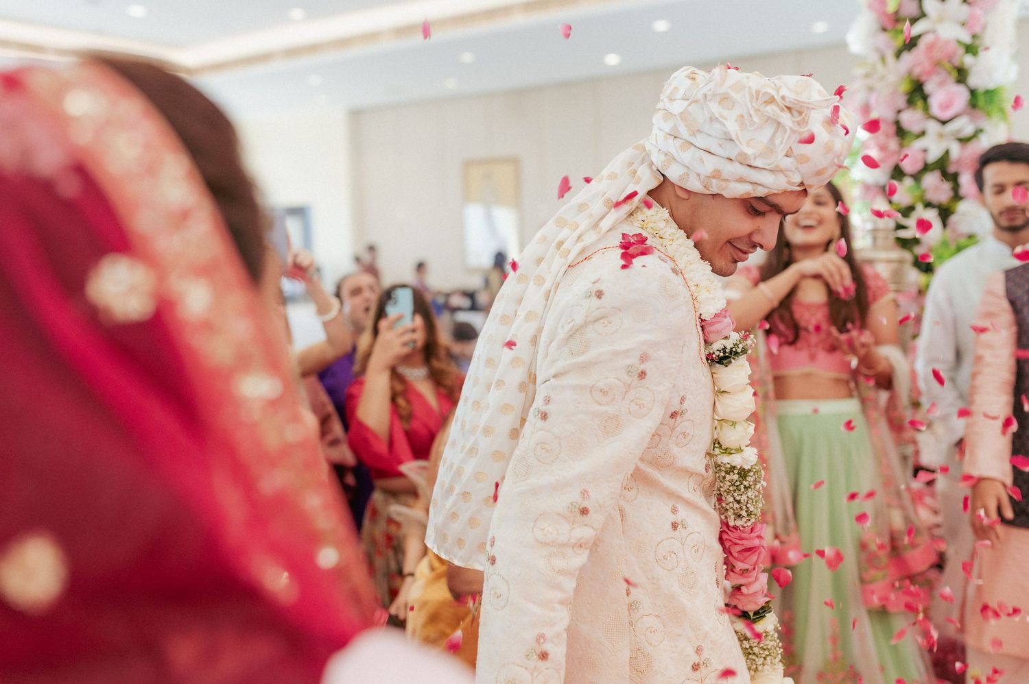 Indian groom in white sherwani laughs as rose petals shower him during vibrant wedding celebration.