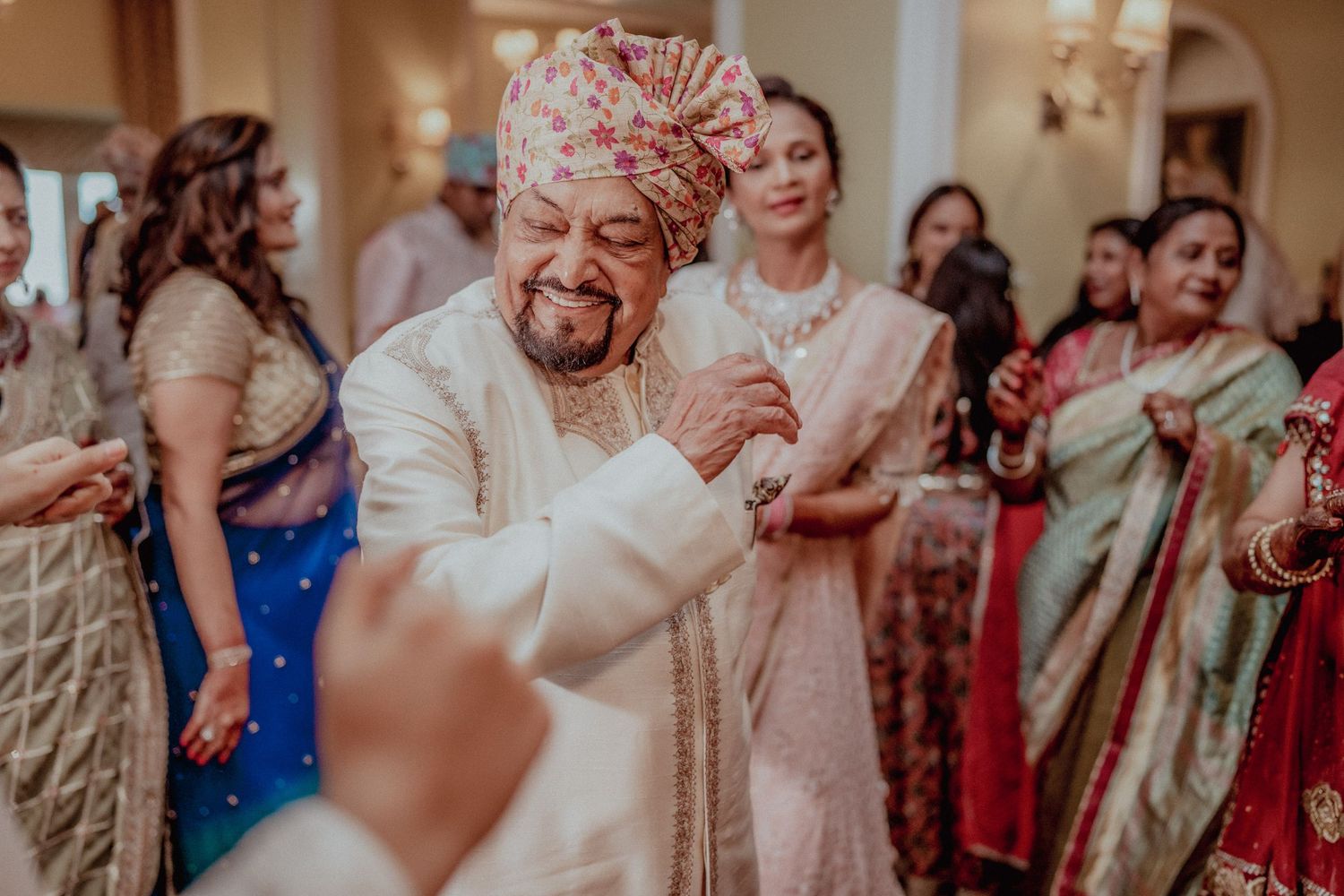 Elderly man in white sherwani dances joyfully surrounded by guests at an Indian wedding.