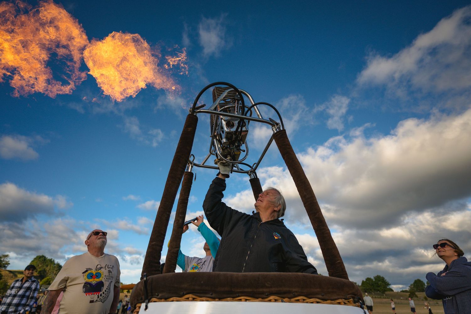Man operates a hot air balloon burner flame against a blue cloudy sky.