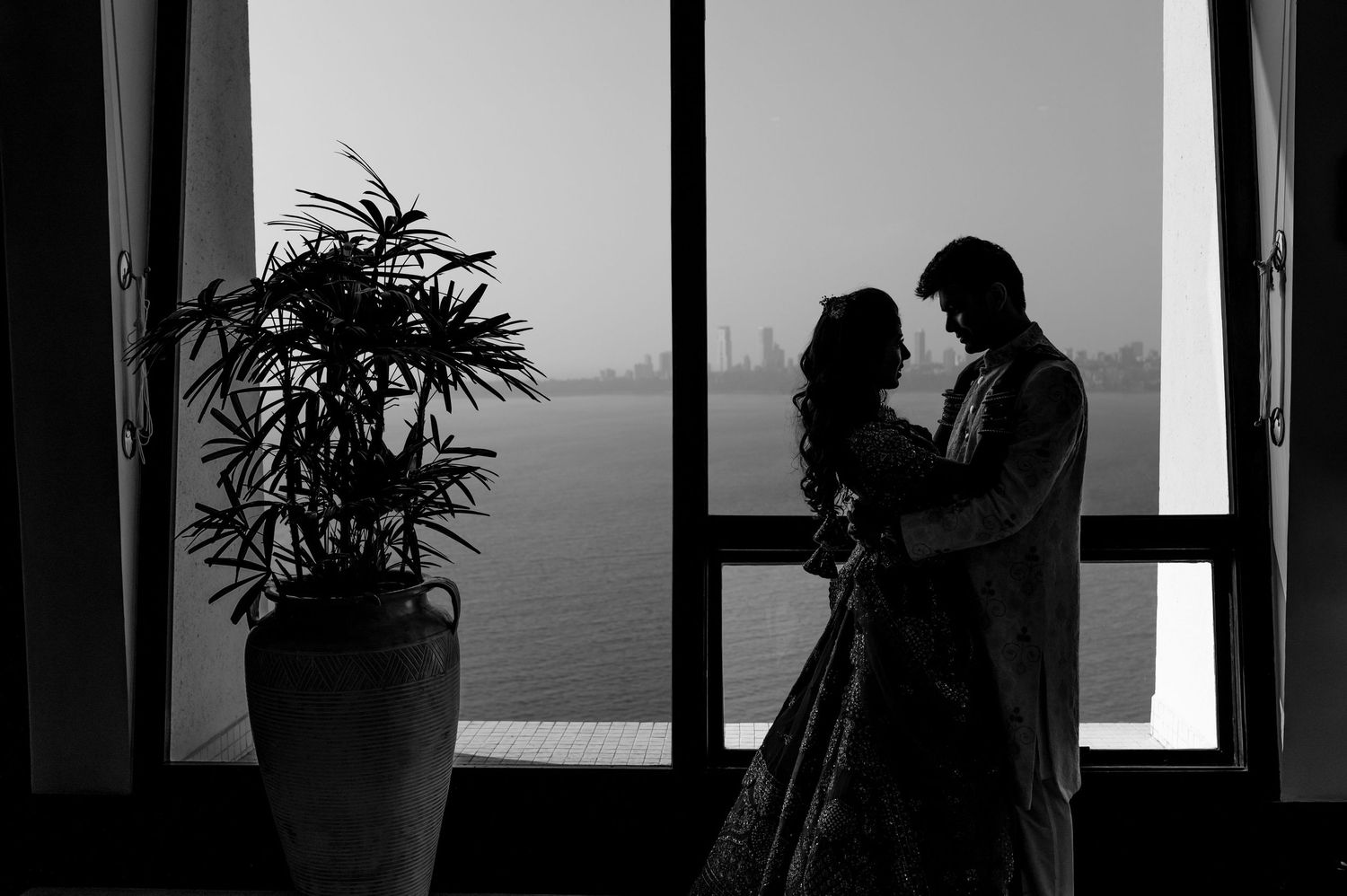 Silhouette of Indian bride and groom embracing by large window with waterfront view in black and white.
