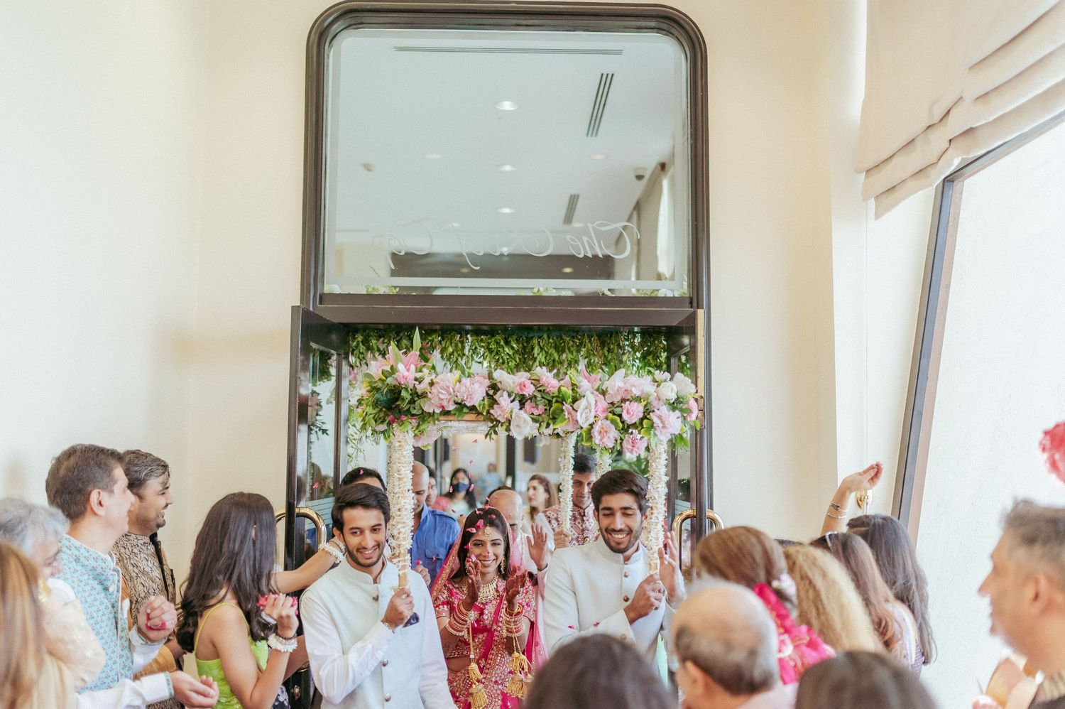 Indian wedding guests celebrate as bride and groom make grand entrance through decorated doorway.