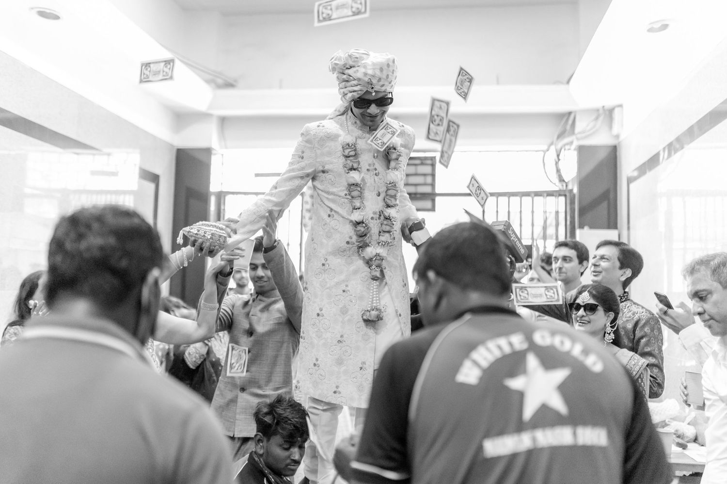 Black and white photo of groom in sherwani lifted by guests at Indian wedding.