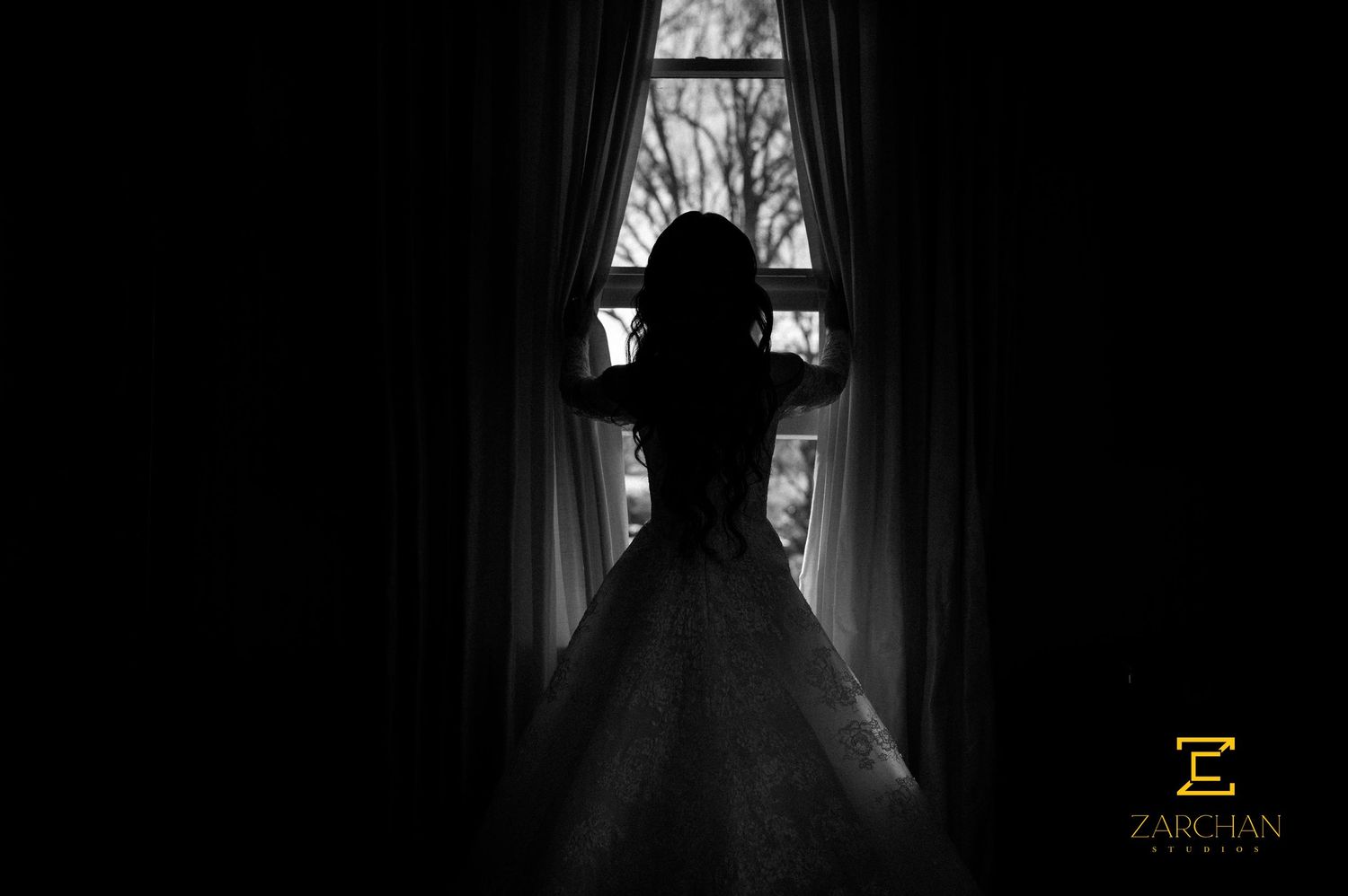 Dramatic silhouette of bride in ball gown standing between curtains at bright window.