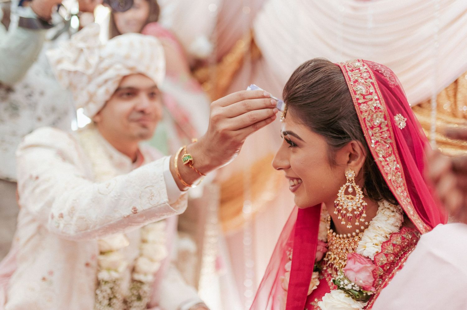 Groom applies sindoor to smiling bride during Hindu wedding ceremony, bride adorned in red lehenga and gold jewelry.