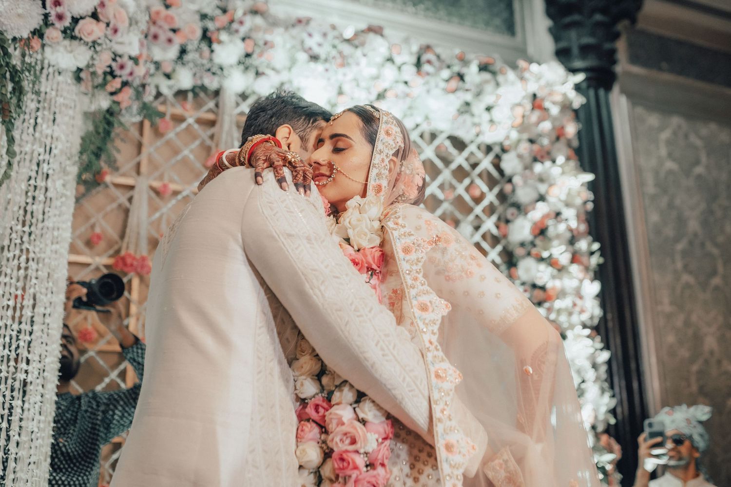Indian bride and groom sharing a tender embrace during their wedding ceremony surrounded by pink flowers.