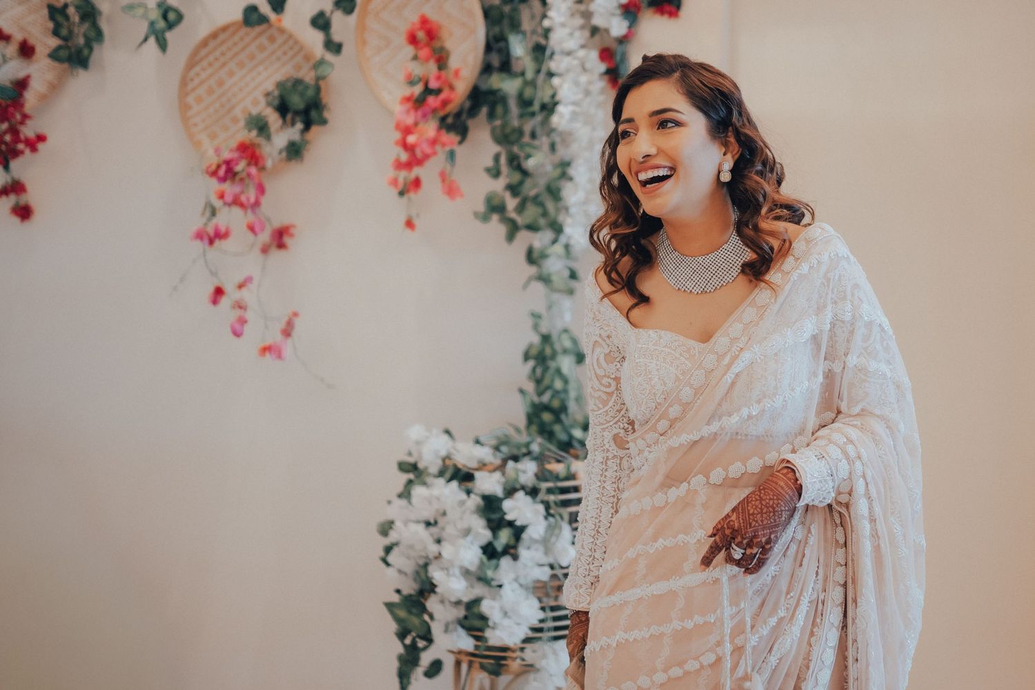 A radiant bride laughs in a white embroidered saree surrounded by floral wedding decor.