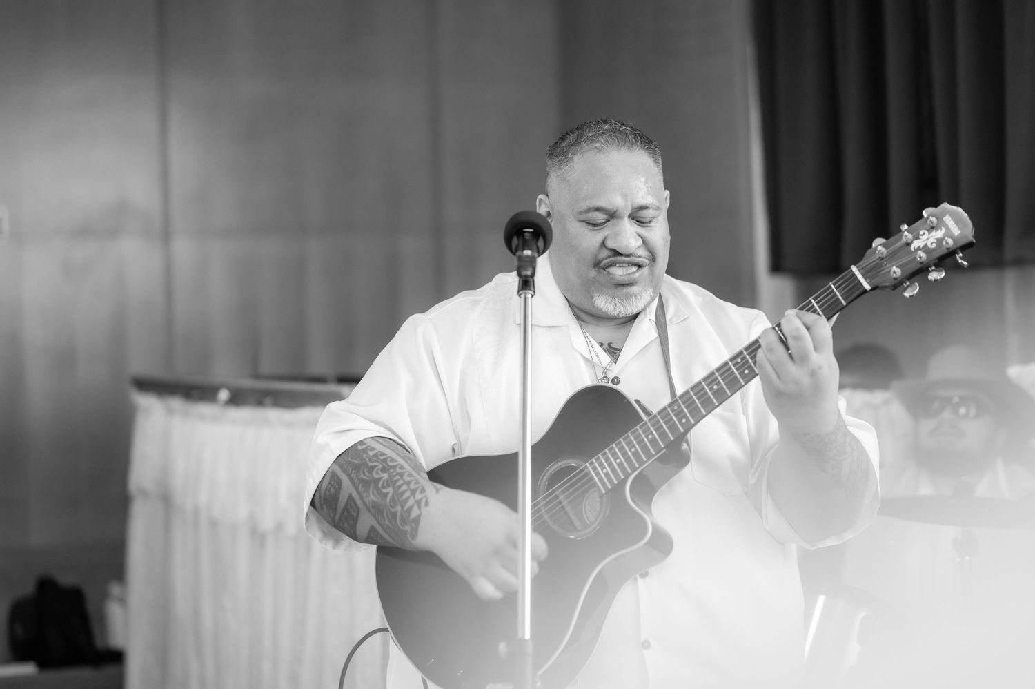 Man playing acoustic guitar and singing into microphone at indoor event in black and white photo.