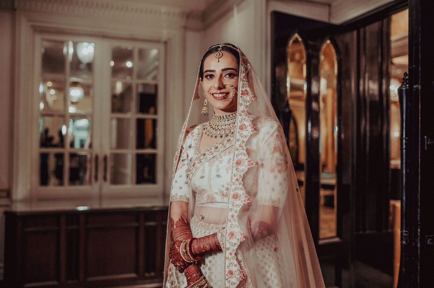 Elegant Indian bride in white and gold lehenga with veil smiling in ornate venue.