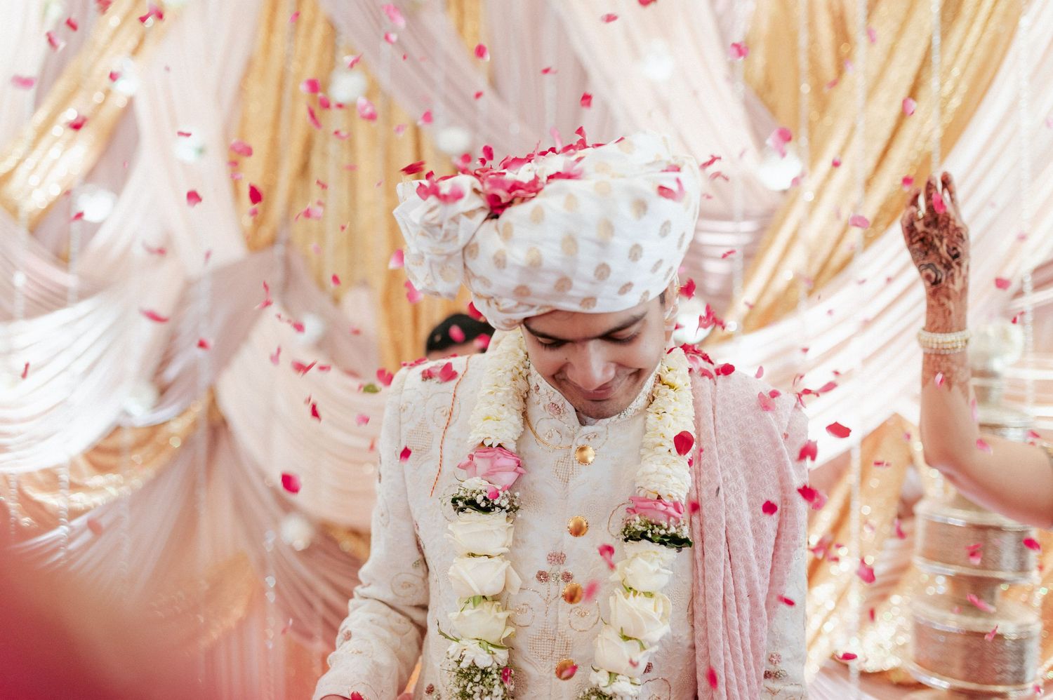 Indian groom in ivory sherwani bows gracefully as colorful flower petals shower during wedding ceremony.