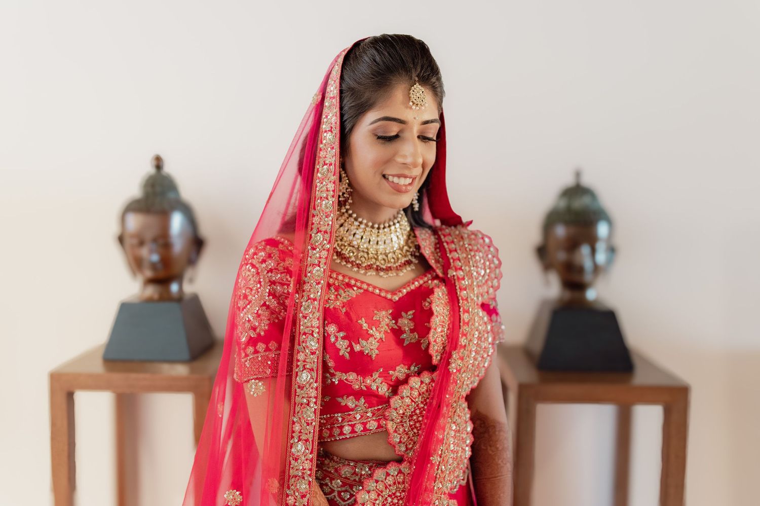 Beautiful Indian bride in red bridal lehenga with gold jewelry smiles softly before her wedding.