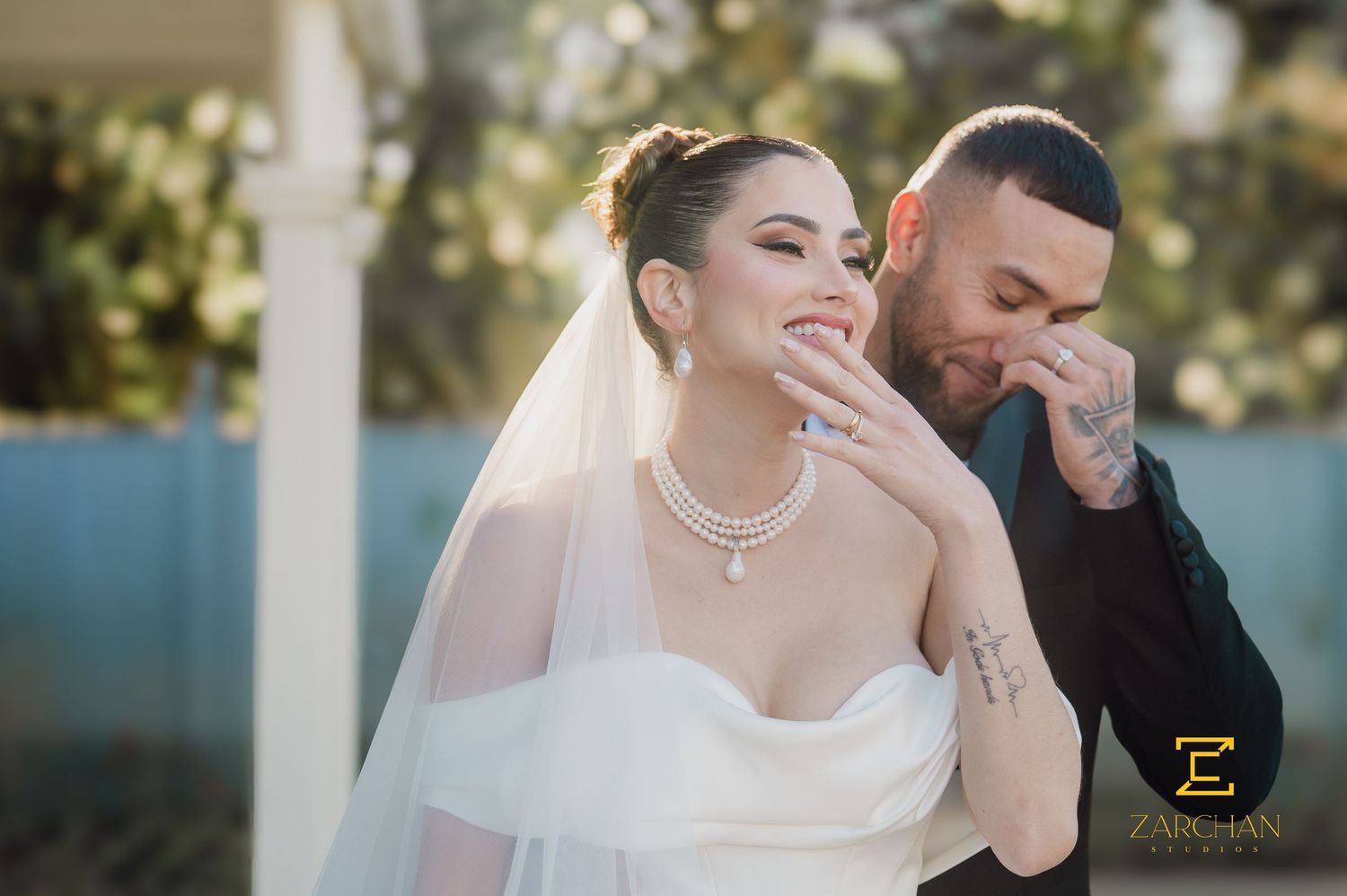 Bride in strapless gown laughs as tattooed groom kisses her hand at outdoor wedding.