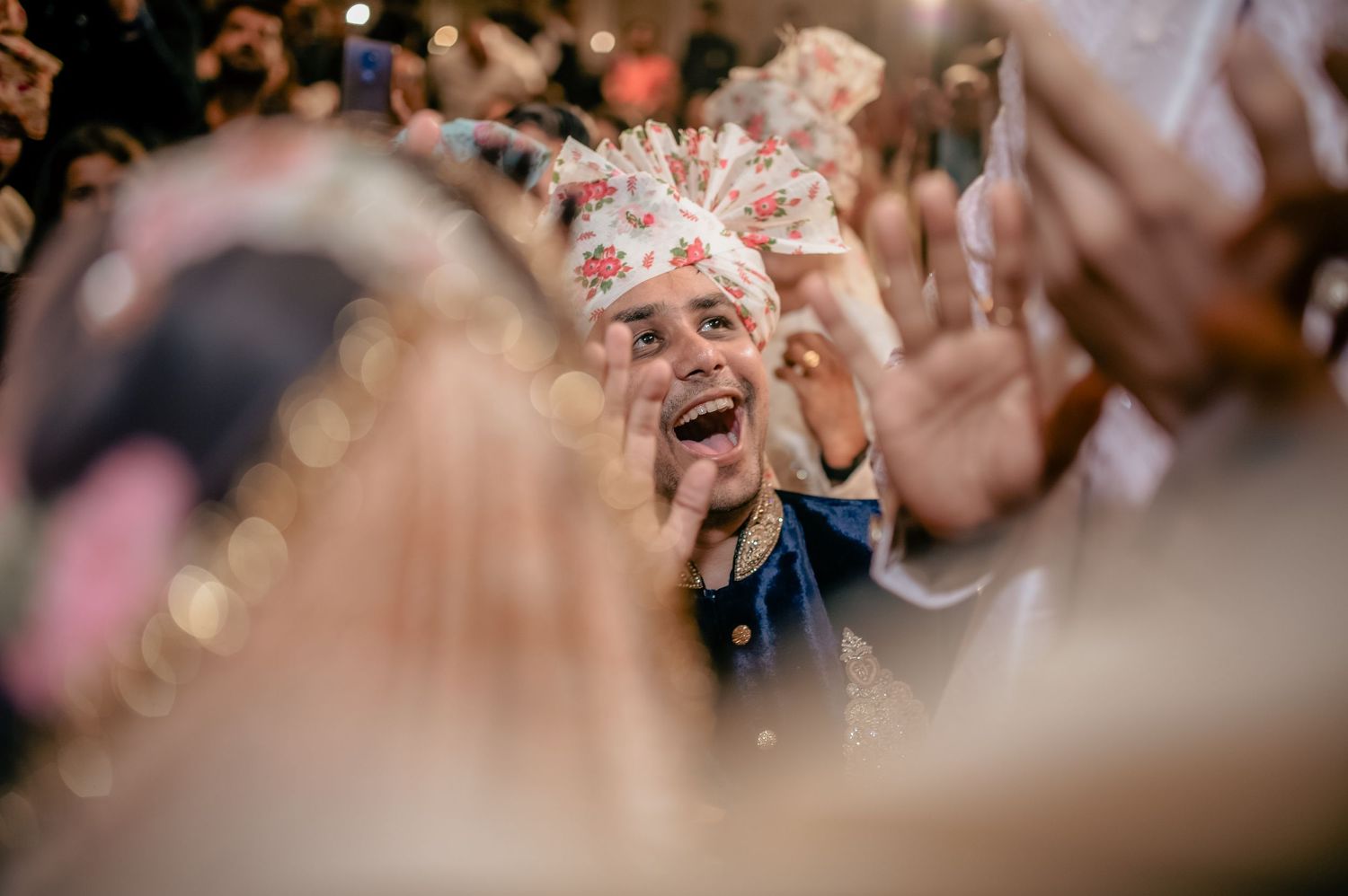 Joyful groom wearing ornate floral wedding crown celebrates surrounded by cheering guests at Indian wedding.