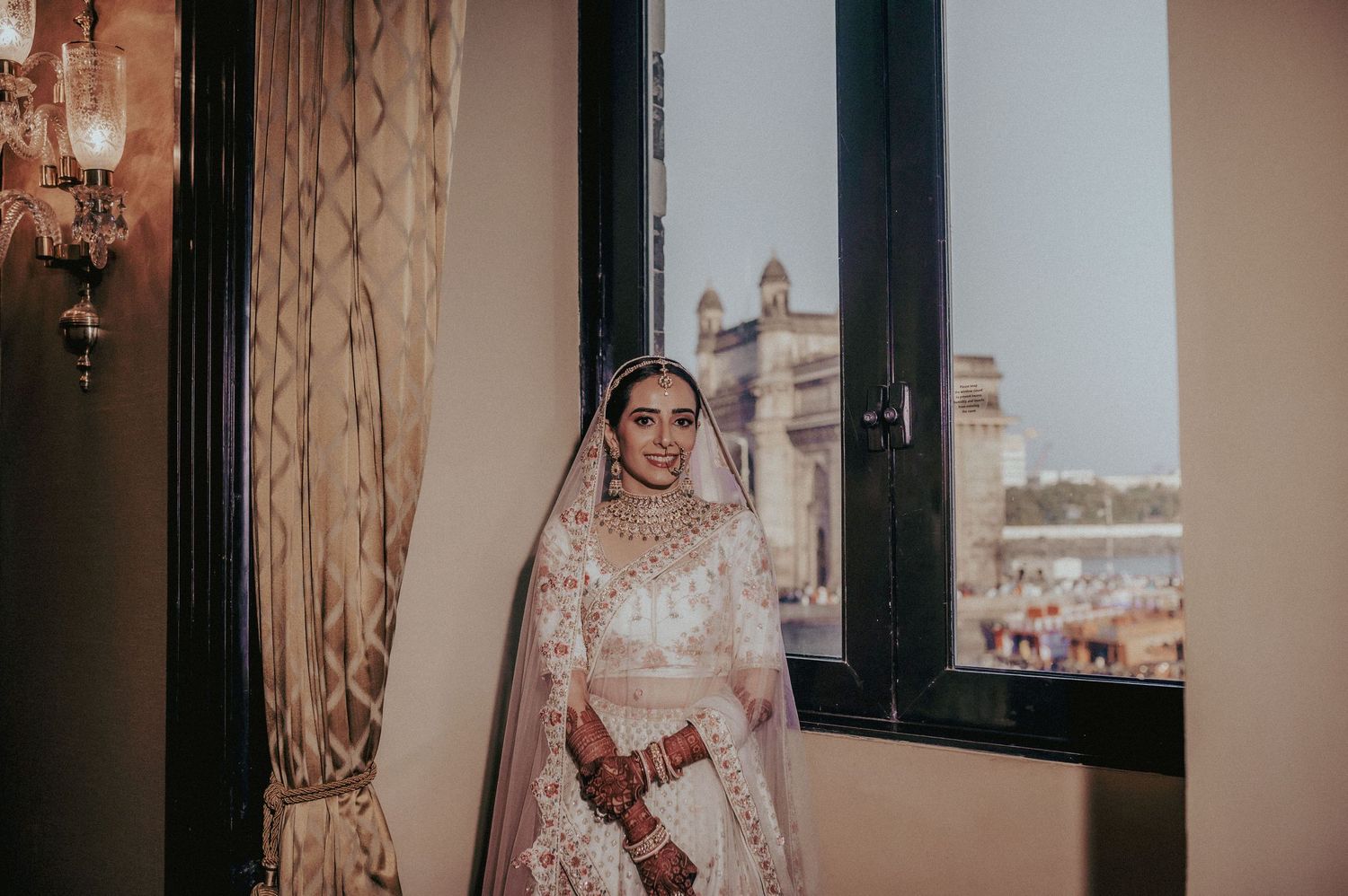 Indian bride in white lehenga standing by window with iconic Gateway of India visible in background.