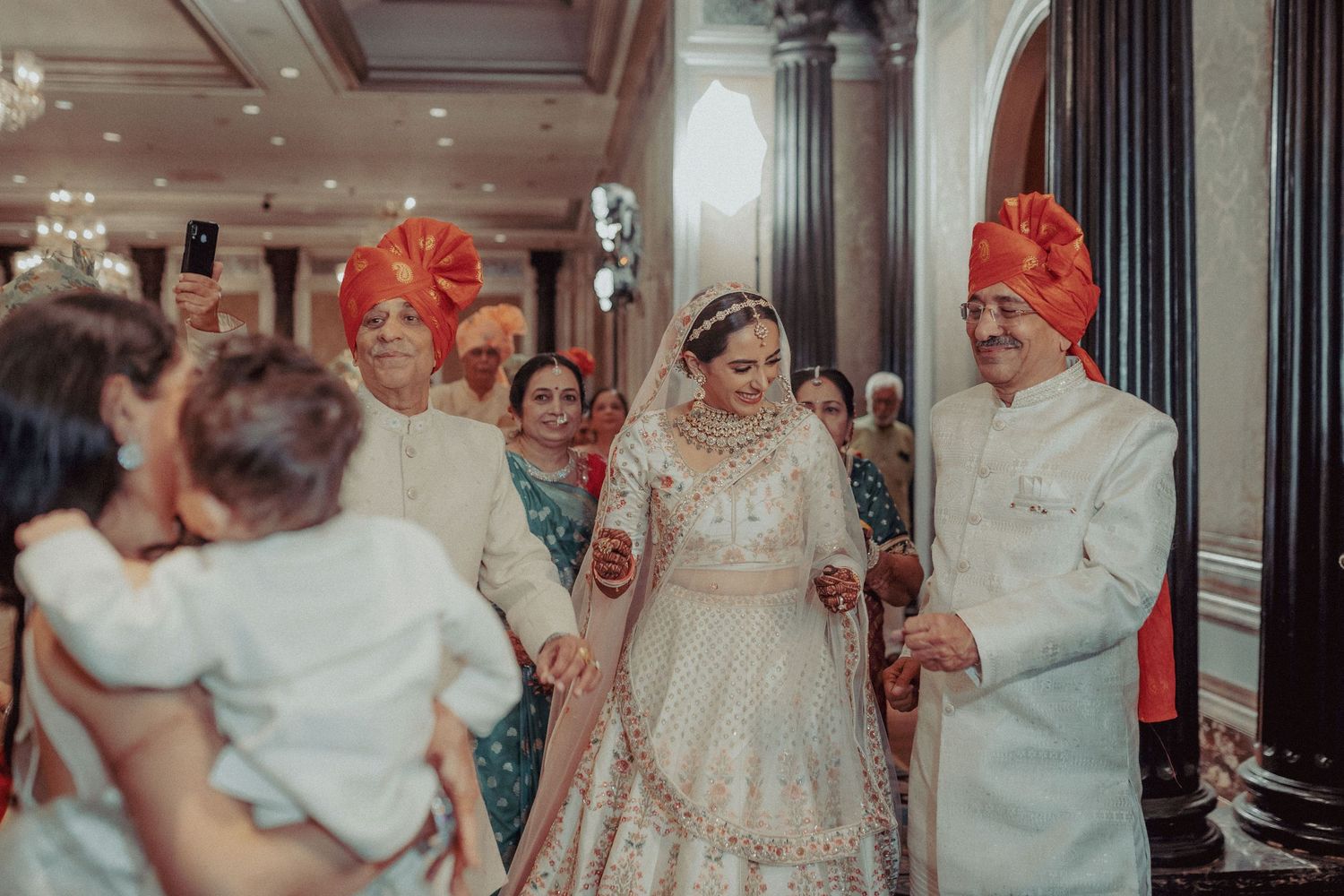 Indian bride in white lehenga escorted by family members in orange turbans at wedding.