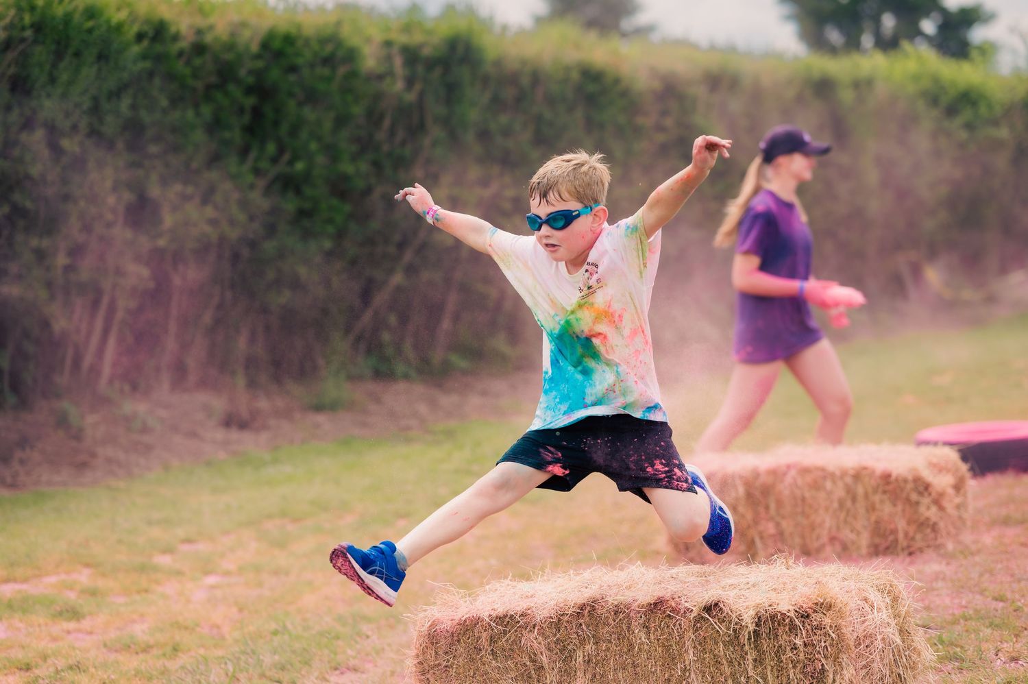 Boy in sunglasses leaping over hay bale during colorful color run race event outdoors.