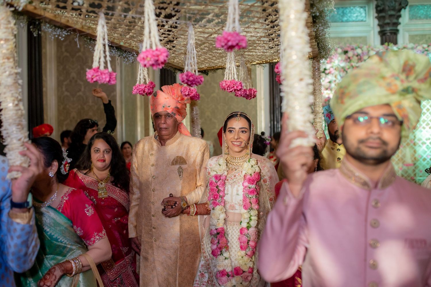 Indian bride in white lehenga processing with family under hanging floral decorations at wedding venue.