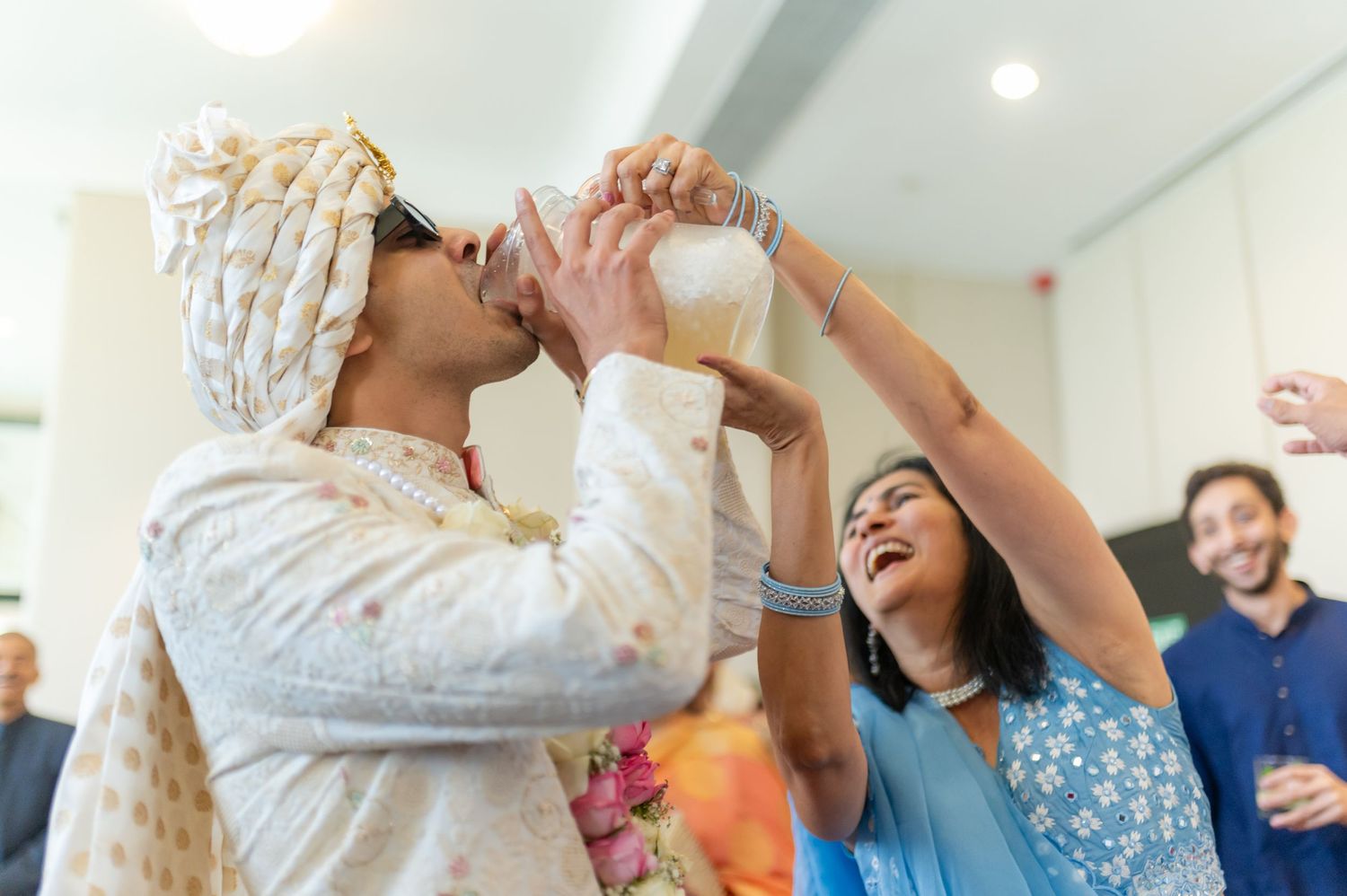 Groom in turban drinks from coconut shell during Indian wedding ceremony while guests celebrate.