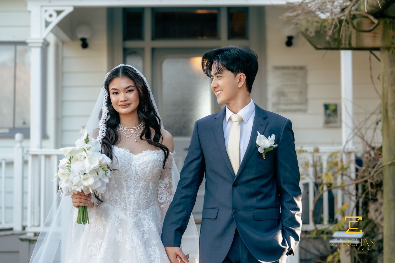 Asian bride and groom walk hand in hand in wedding attire outside a white Victorian home.