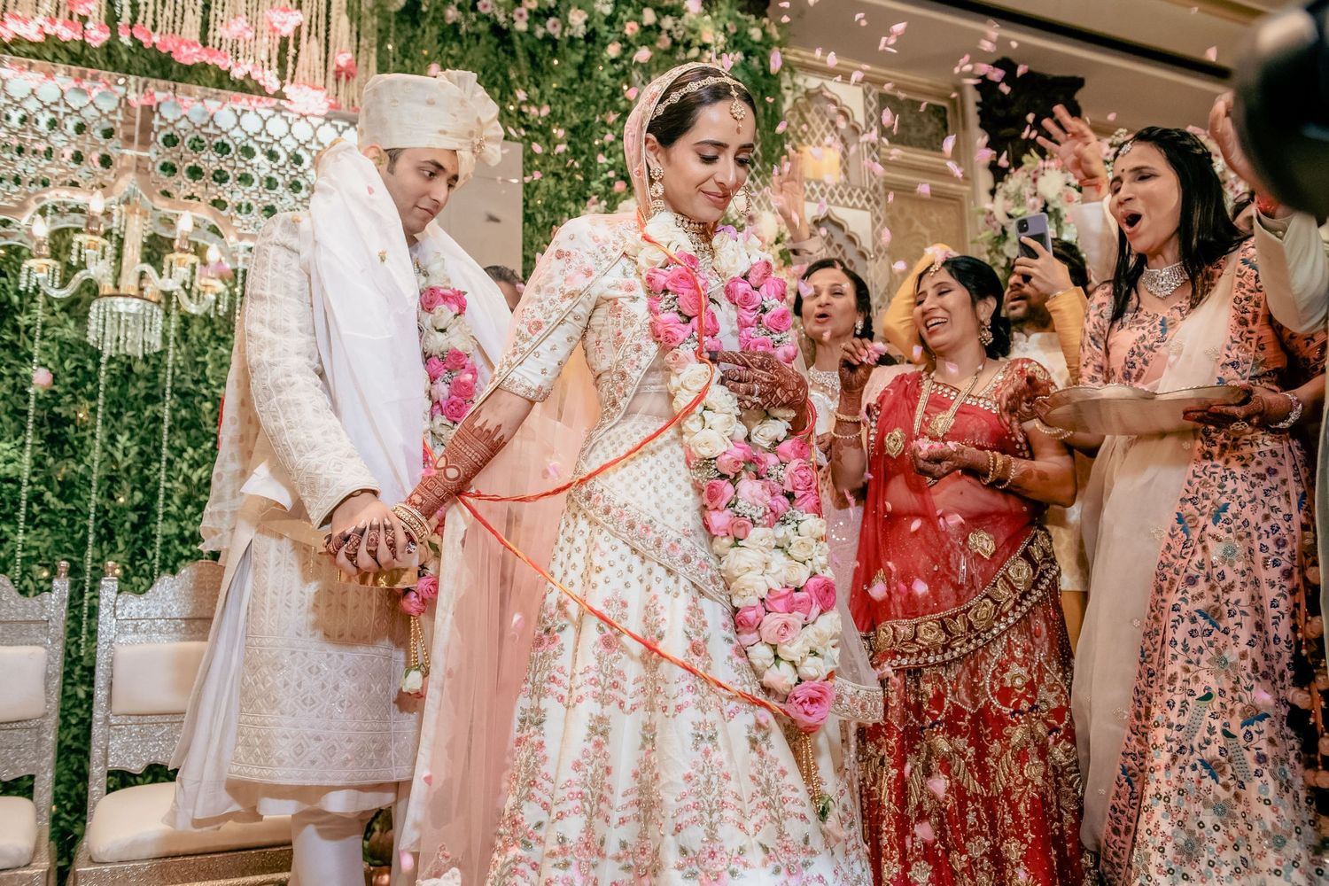 Indian bride in white lehenga walks with groom amid celebrating guests at wedding.
