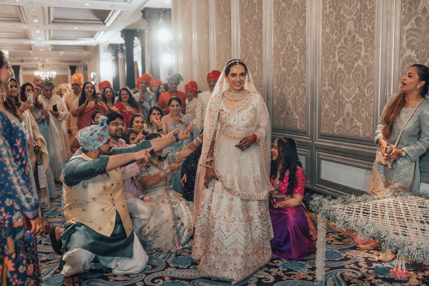 Indian bride in white lehenga walking through guests during traditional wedding ceremony in grand hall.