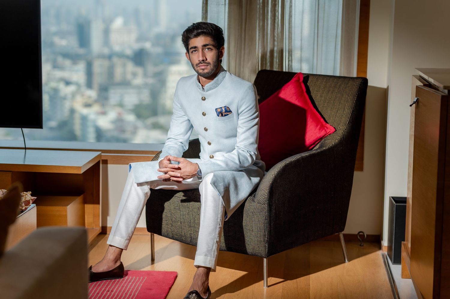 Groom in white sherwani sitting in chair with city skyline view.