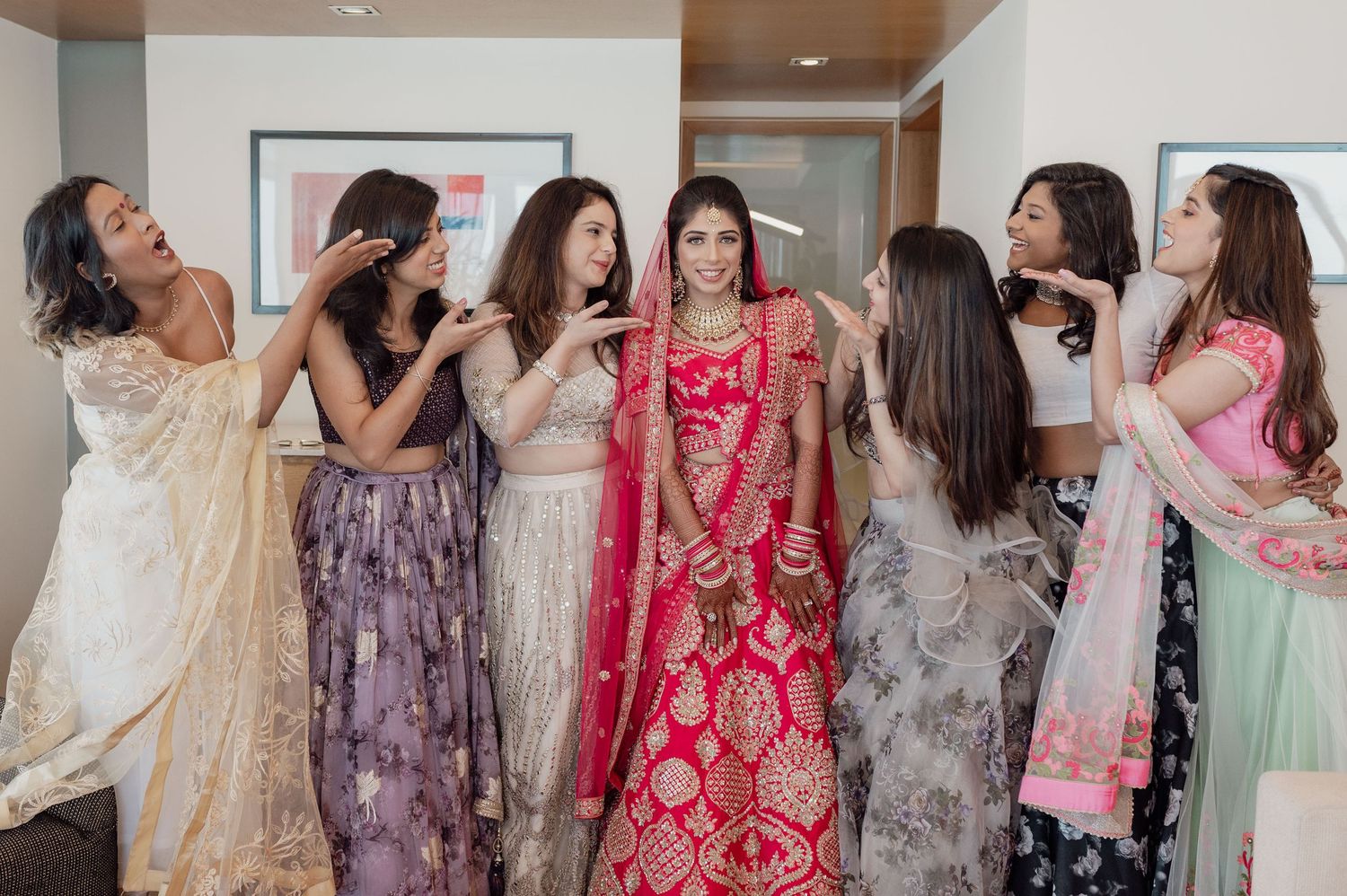 Indian bride in red lehenga surrounded by bridesmaids in colorful attire at wedding preparation.