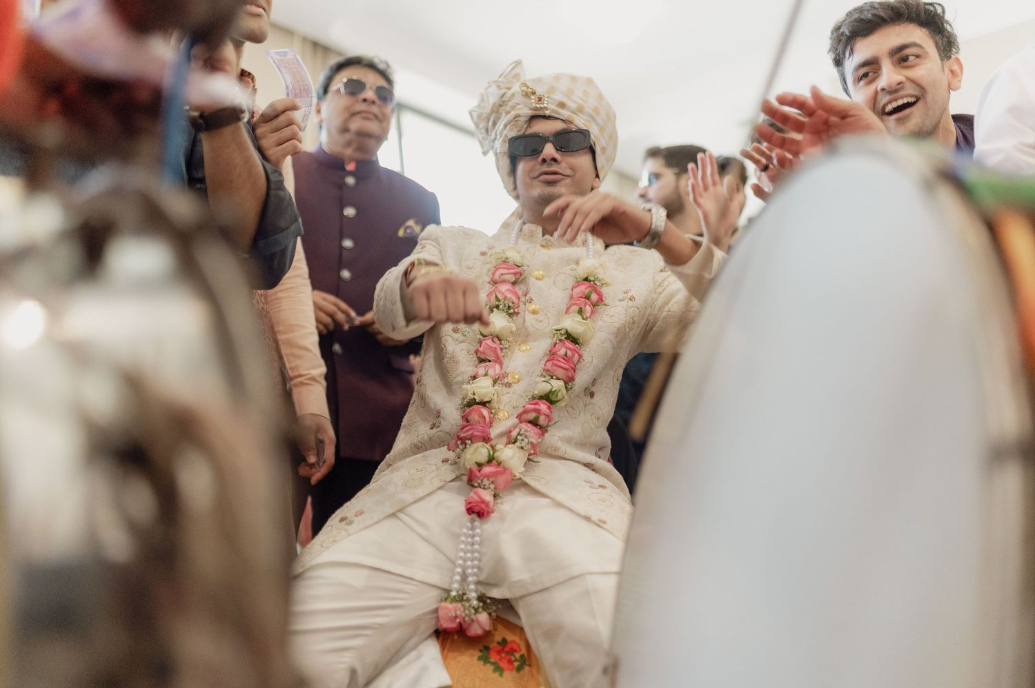 Groom in white sherwani and sunglasses dances joyfully during Indian wedding baraat procession.