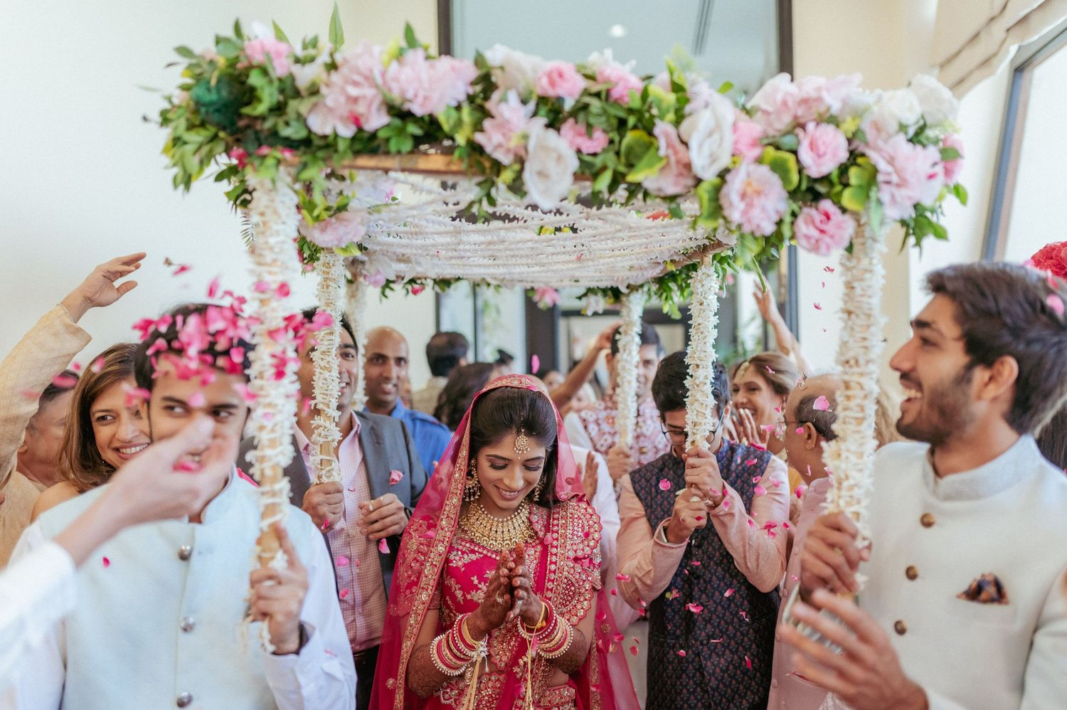 Indian bride in red lehenga walks under pink floral canopy surrounded by joyful wedding guests.