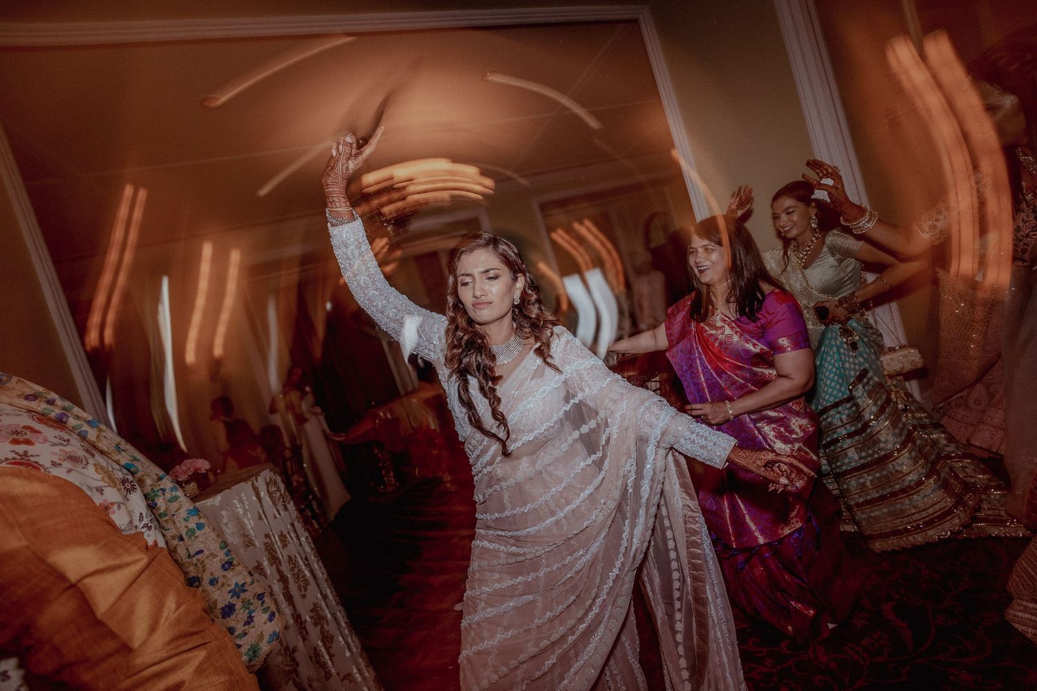 Bride in white lehenga dancing joyfully at Indian wedding celebration with guests in colorful sarees.