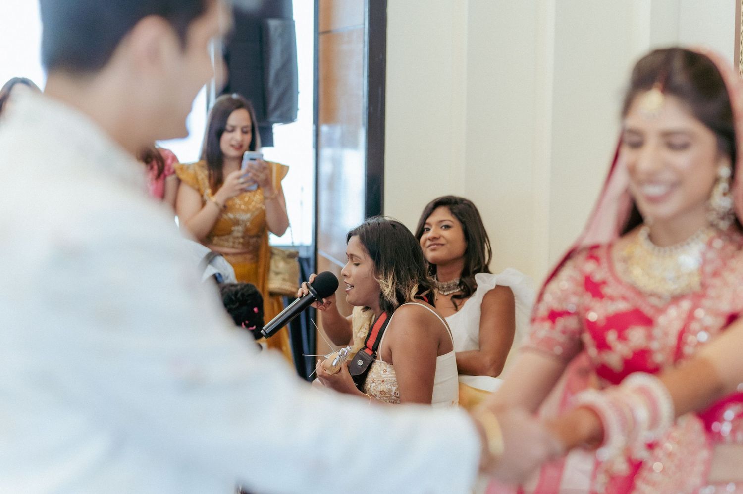 Indian bride in red lehenga smiling as guests perform live music at wedding ceremony.