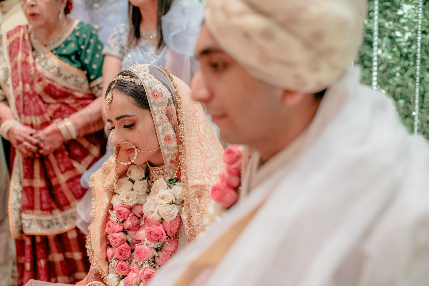Bride in peach lehenga with floral garland bows head during traditional Indian wedding ceremony.