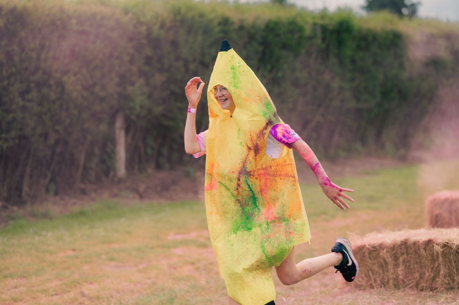 Person in yellow banana costume jumping over hay bale covered in colorful powder at color run.