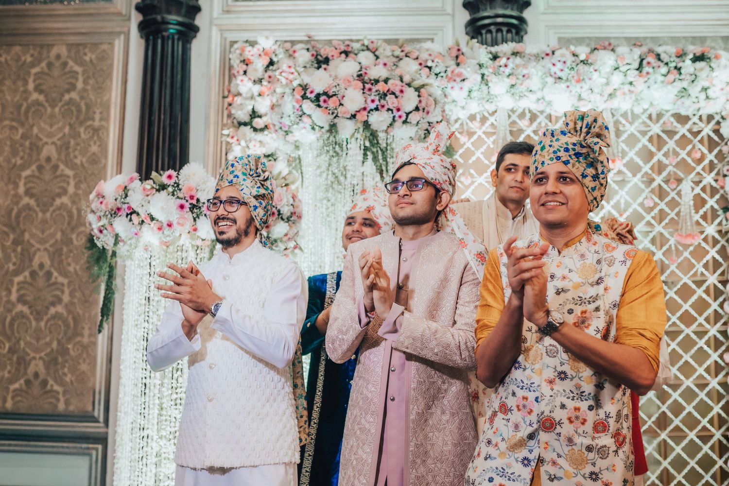 Groom and groomsmen in colorful turbans clapping joyfully at Indian wedding ceremony with floral decor.