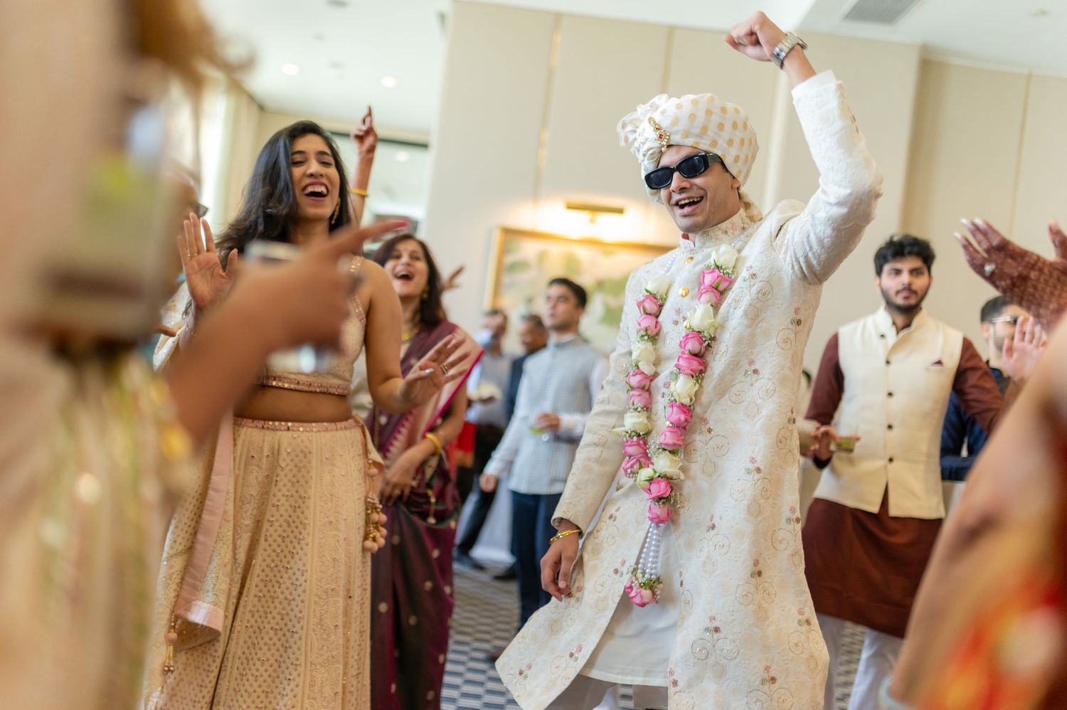 Groom in white sherwani with sunglasses dancing joyfully at baraat ceremony.