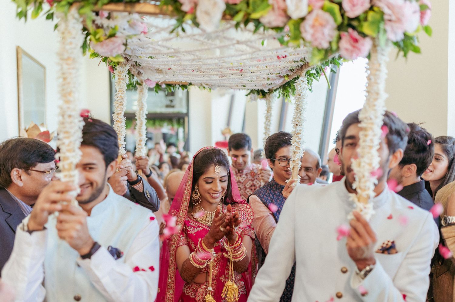 Indian bride in red lehenga walks under floral canopy surrounded by family at traditional wedding.