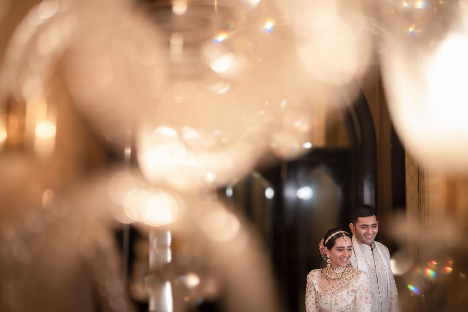 Indian wedding couple stands romantically beneath glowing bokeh lights and elegant crystal chandeliers.