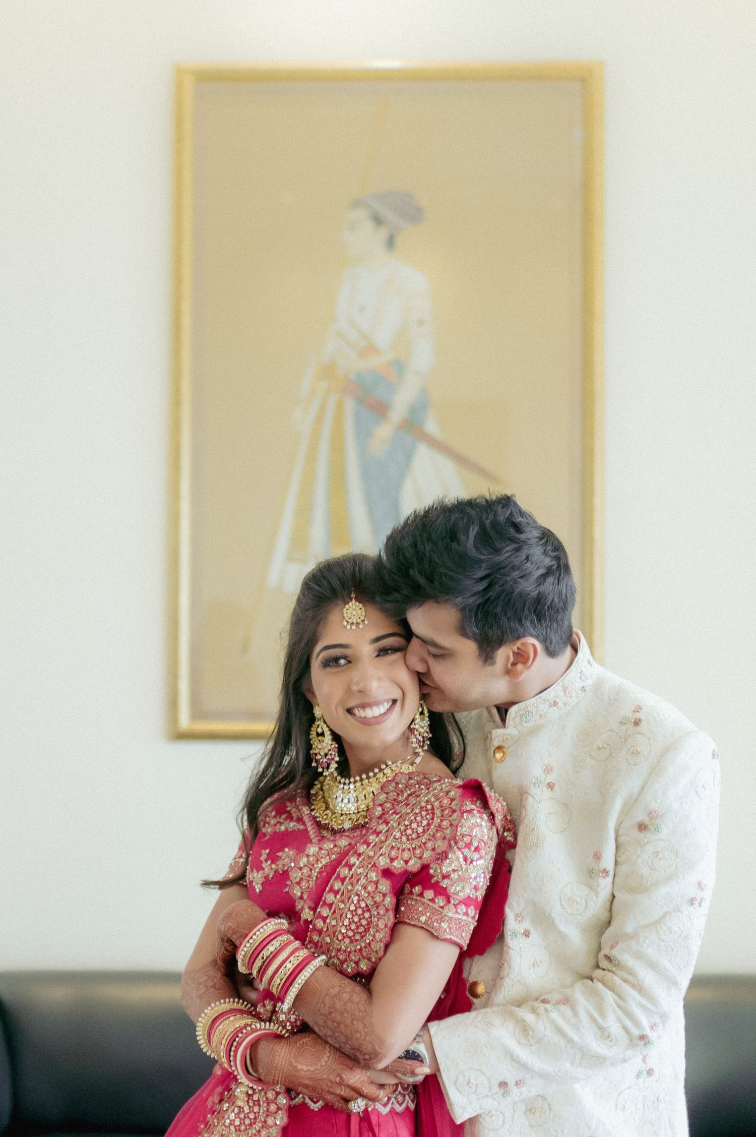 Indian bride in red lehenga with gold jewelry receives a kiss from groom in white sherwani.