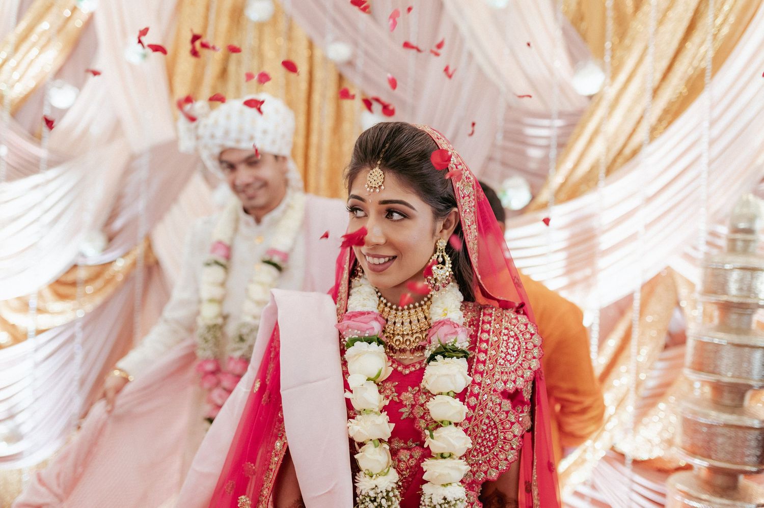 Radiant Indian bride in red lehenga and floral garland smiles during traditional Hindu wedding ceremony.