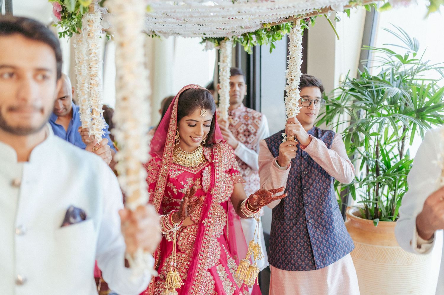 Bride in red lehenga walks under floral canopy during traditional Indian bridal entry.