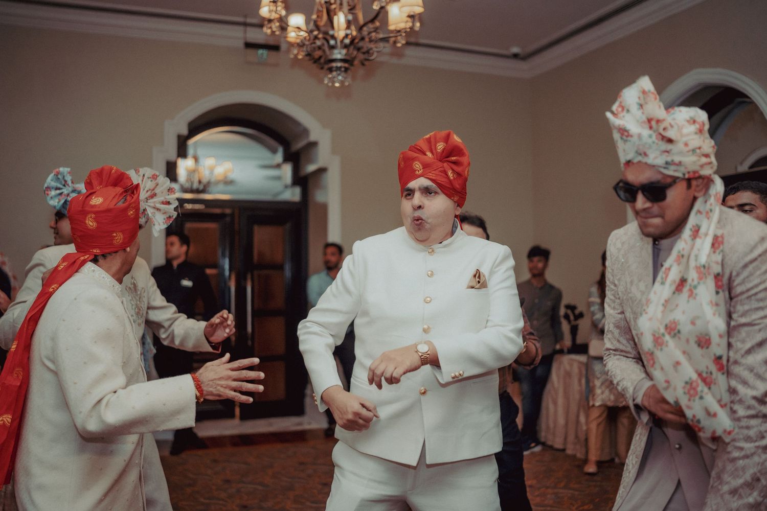 Indian groom in white sherwani dancing with groomsmen in orange turbans at wedding reception.