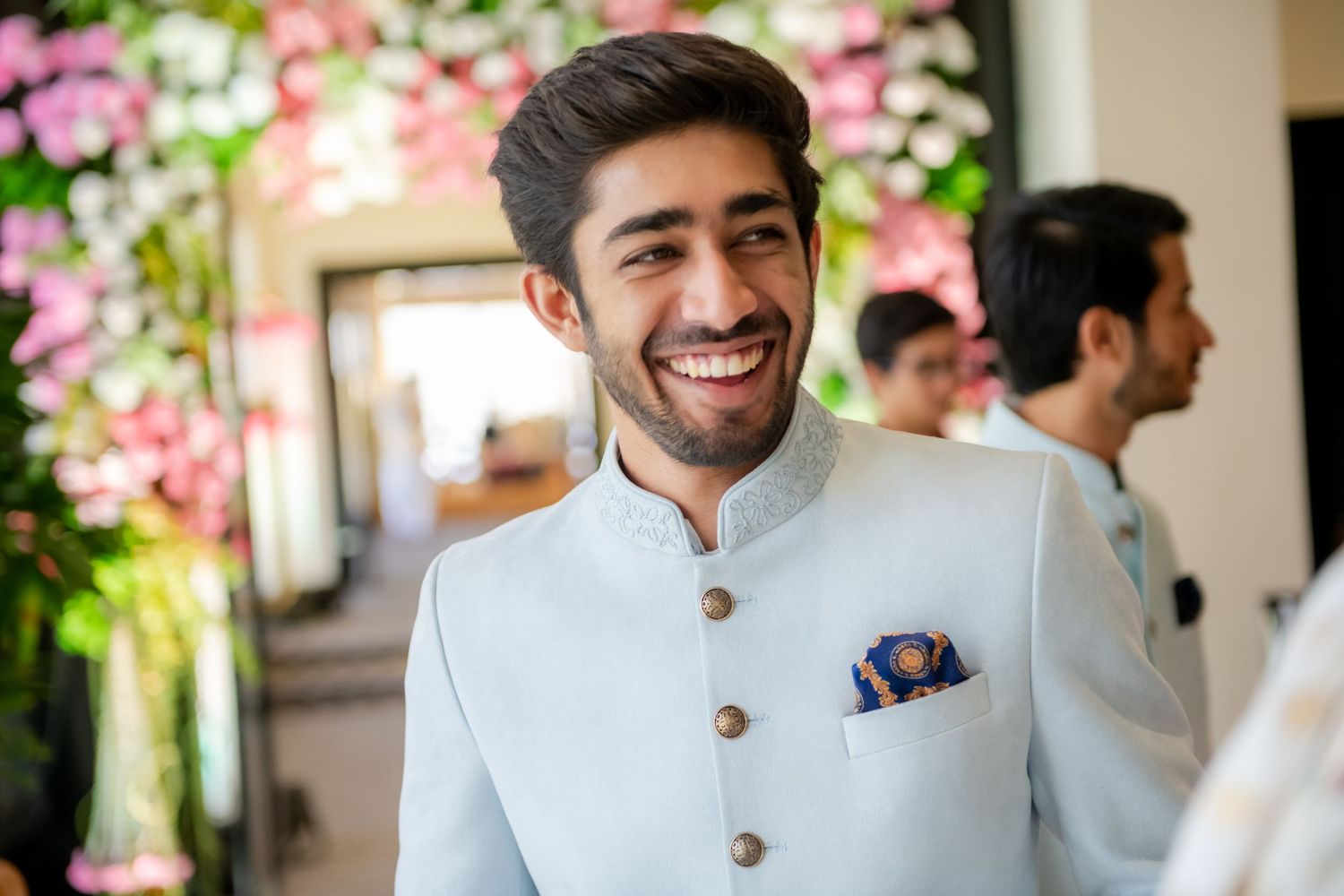Smiling groom in light blue sherwani with floral backdrop at wedding.