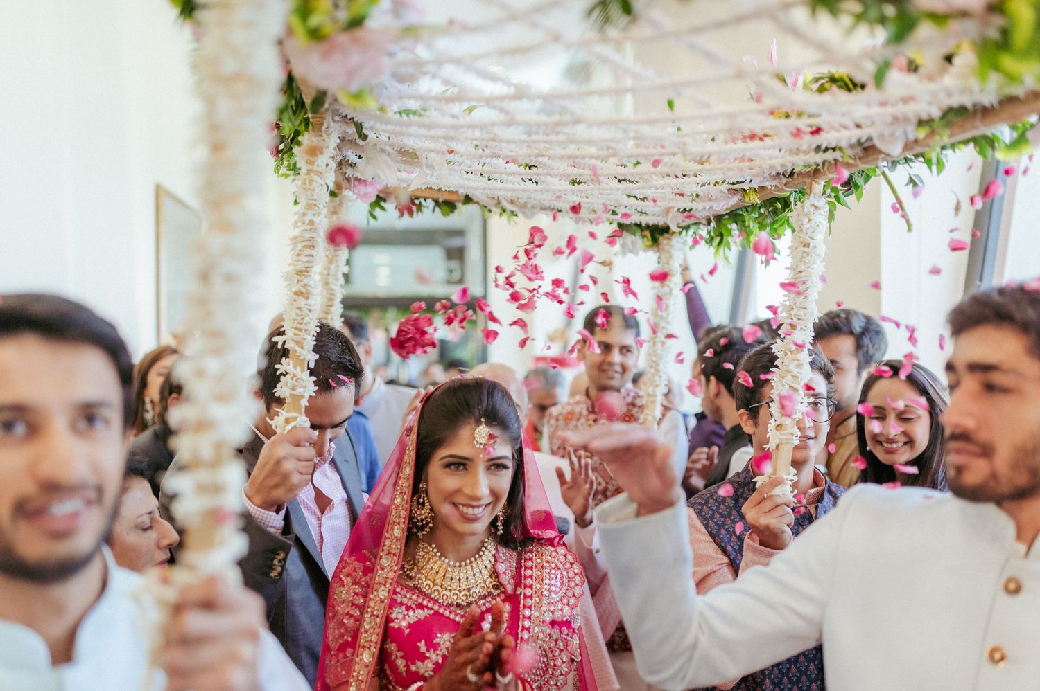 Bride in vibrant red bridal lehenga walks under decorated floral mandap with joyful wedding guests.