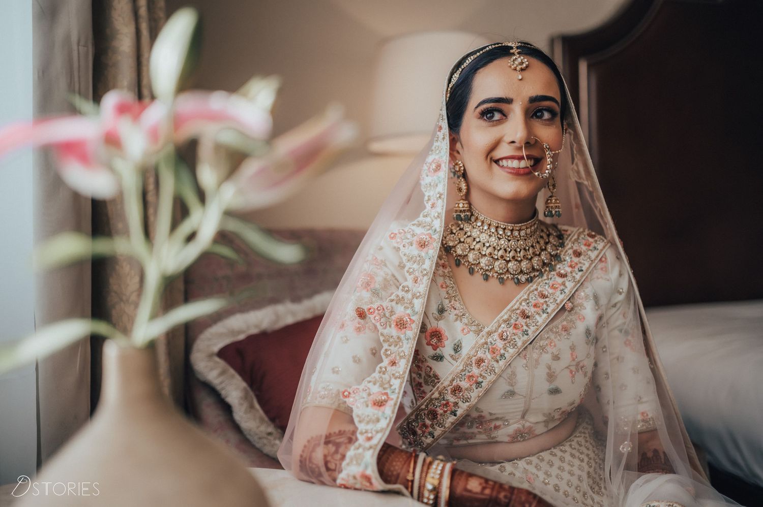 Beautiful Indian bride wearing white floral lehenga with gold necklace and dupatta smiles gracefully.