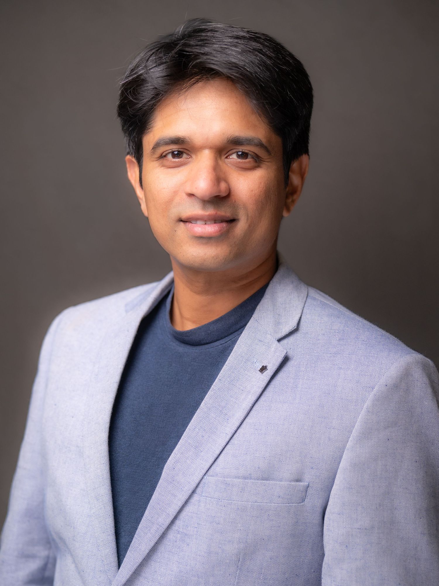 Professional headshot of a man in a light grey blazer and navy shirt against a grey studio backdrop.